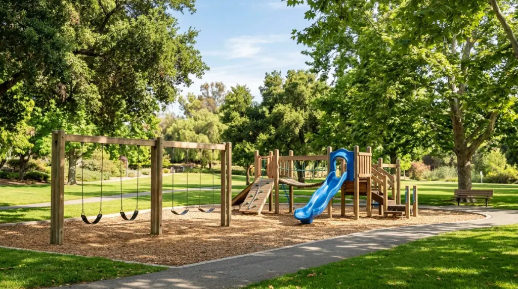 A tranquil scene of a park playground with swings and a slide, surrounded by mature shade trees, on a sunny afternoon.