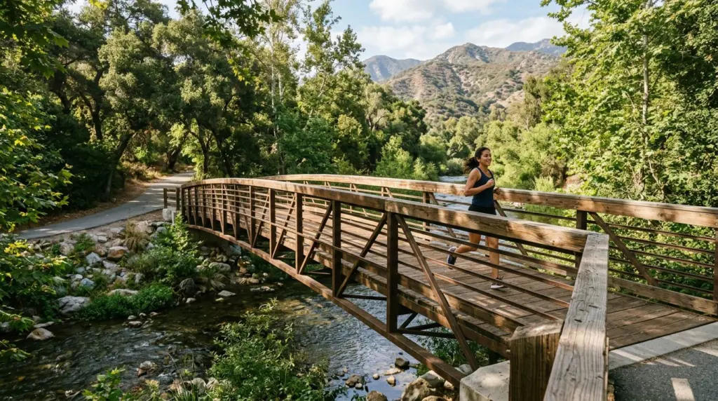 A well-maintained pedestrian bridge over the San Gabriel River Trail in Azusa, with dappled sunlight filtering through overhead trees and the river flowing gently below.